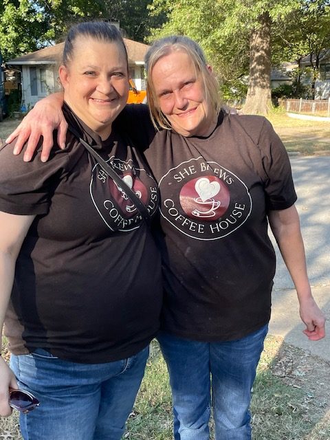 Two female co-workers in matching uniform t-shirts side-hugging while smiling at the camera