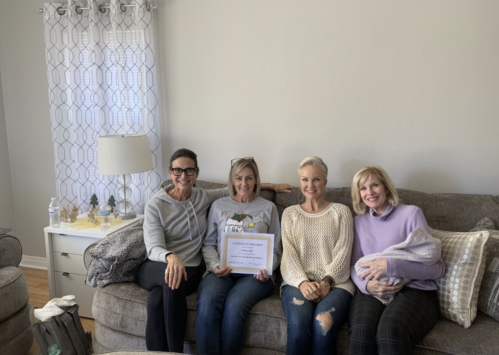four women sitting on a couch together smiling while one holds up a certificate